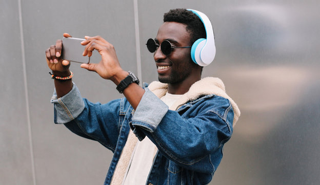 Modern happy smiling young african man taking selfie picture by smartphone while listening to music in wireless headphones on gray wall background