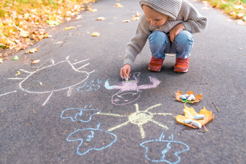 Naklejka premium little boy of 3-5 years old, sits on pavement and draws drawings with colorful crayons, free space for creativity, children. In warm clothes on autumn day nature.