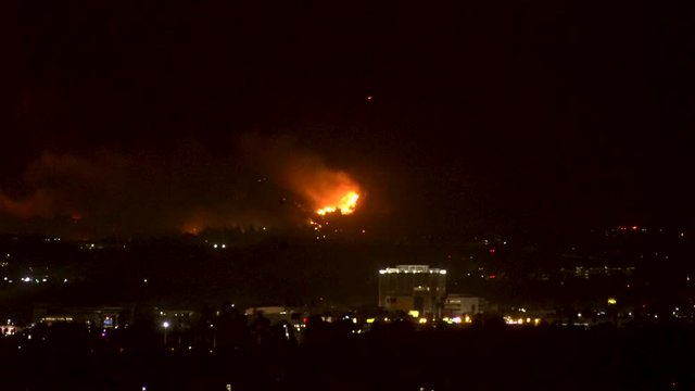 Forest fire on mountain range above residential neighborhood at night as planes dump water and smoke spreads helicopters fly around with search lights.