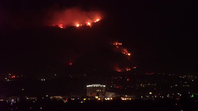 Forest fire on mountain range above residential neighborhood at night as planes dump water and smoke spreads helicopters fly around with search lights.