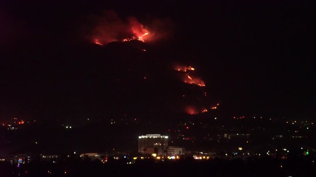 Forest fire on mountain range above residential neighborhood at night as planes dump water and smoke spreads helicopters fly around with search lights.
