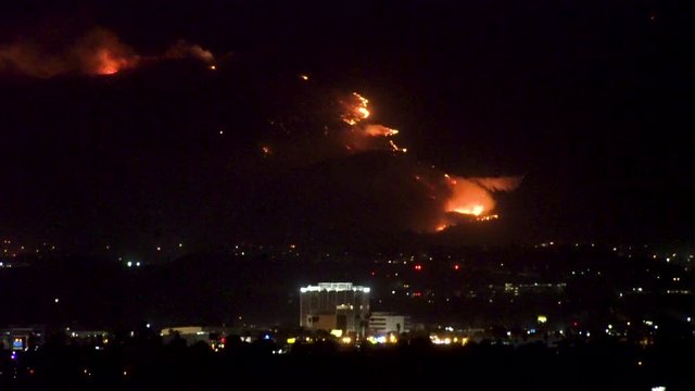 Forest fire on mountain range above residential neighborhood at night as planes dump water and smoke spreads helicopters fly around with search lights.