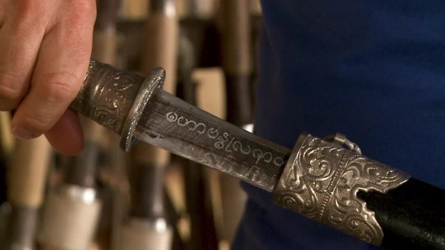 A daylight extreme closeup shot of a local vendor removing a traditional Thai sword made from forged metal from its wooden sheath casing decorated with brass fittings..