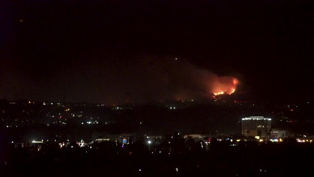 Forest fire on mountain range above residential neighborhood at night as planes dump water and smoke spreads helicopters fly around with search lights.