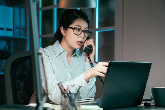 Young asian japanese businesswoman talking on phone and using laptop computer at office desk late night in front of windows overlooking city. lady worker having problem on project discuss colleague