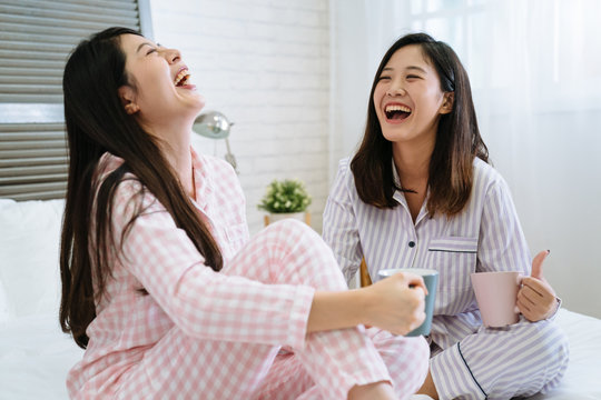 Close up lifestyle indoor fresh portrait of two pretty smiling friends. asian chinese girls sitting in cozy white bed wearing pink and purple pajama enjoy good time. female cheerful laughing chatting