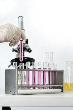 Hand In White Glove Holding Glass Test Tube With Purple Violet Liquid Water Close Up With Test Tubes And Microscope On White Background, Cosmetic Science Laboratory Research Study Education Concept.