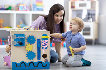 Cute toddler baby and his mom playing with busy board at home or daycare. Children educational toys. Wooden game diy board