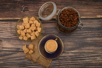 A Cup of coffee, coffee beans in a glass jar and pieces of brown sugar on a wooden background.