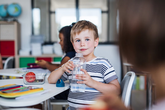 Small School Boy Sitting At The Desk In Classroom, Drinking Water.