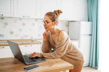 Beautiful smiling young woman fair long hair girl in glasses wearing in cozy knitted sweater using laptop at bright kitchen