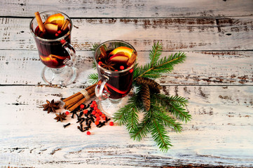 Two glasses of mulled wine with spices and fruit slices stand on a table surrounded by fir branches...