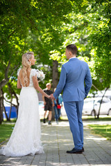Fototapeta premium Young beautiful couple of newlyweds bride and groom walk holding hands in a park with green trees on their wedding day. Love and lovestory of wife and husband before starting a family.