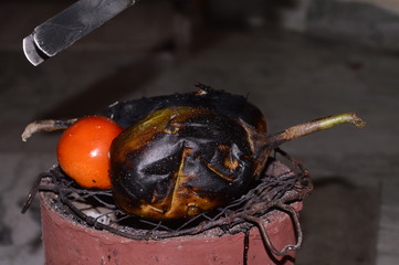 close up of Grilling of Brinjal and tomato on coal stove or charcoal stove ( or mitti ka chulha or charcoal chulha or coal chulha or charcoal shegadi ) in traditional style