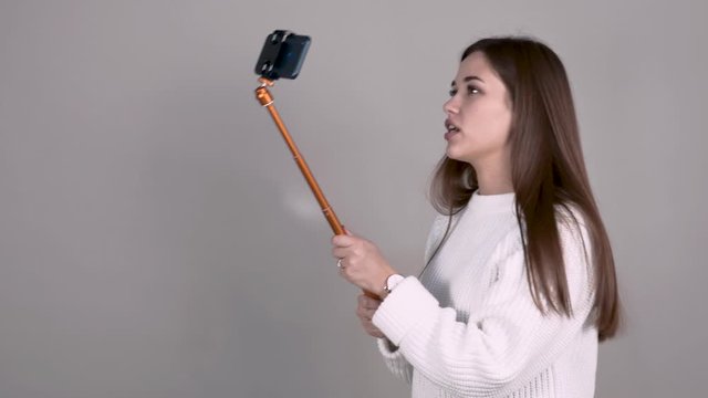 caucasian Girl author of video blog brown haired white jumper long hair on light gray background holds selfiestick withsmartphone and emotionally communicates with her subscribers leads live broadcast