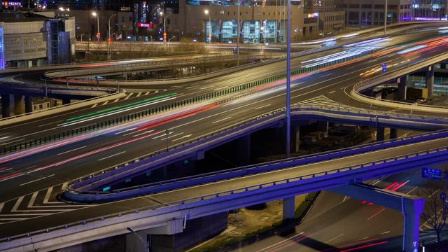 Timelapse Beijing automobiles drive in heavy traffic along illuminated Chinese overpass road interchange against city district buildings at night