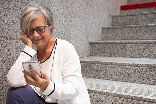 A Pretty Woman Dressed In White Sitting On The Stairs Of A Shopping Mall Looking At Her Cell Phone. A 70 Year Old Female With Gray Hair And Glasses.