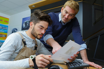 mature mechanic helping younger colleague sat at computer desk