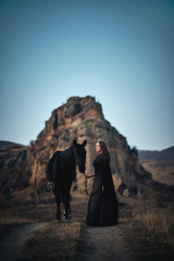 a lovely young girl in a black light dress stands in a field at sunset with her black horse on a background of mountains