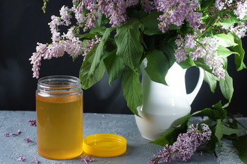An open jar of linden honey stands on a table next to a bouquet of purple lilac in a white jug
