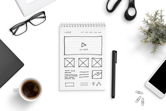 Sketch of a website on a paper writing pad, surrounded by smart phone, cup of coffee, glasess, plant. Office desk, top view, flat lay.