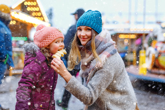 Young mother and daughter eating white chocolate covered fruits on skewer on traditional German Christmas market. Happy girl and woman on traditional family market in Germany, Munich during snowy day.
