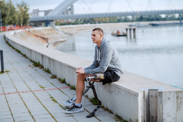 Handsome sporty caucasian handicapped man in sportswear and with artificial leg sitting on quay.