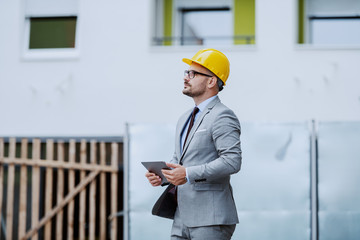 Side view of serious dedicated hardworking architect in gray suit and with yellow helmet on head holding tablet and looking at building while standing at construction site.