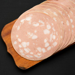 Sliced Mortadella Bologna Meat on a rustic wooden board over black surface, top view. Flat lay, from above, overhead. Close-up. © Liudmyla