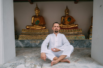 A man sits in a lotus position in a Buddhist palace on the background of the Buddha.
