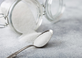 Glass jar of natural white refined sugar with silver spoon on light table background.