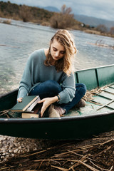 Fototapeta premium Portrait of young beautiful woman sitting on a boat and reading a book. She is bookworm and she choose between few books. 