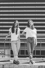 Black and white shot of Two young hipster women leaning back on fence and making fun in front of urban wall
