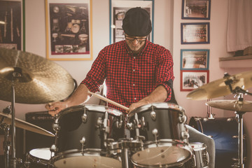Caucasian young man with glasses and beret wearing a red and black plaid shirt, playing the drums with a soft side light