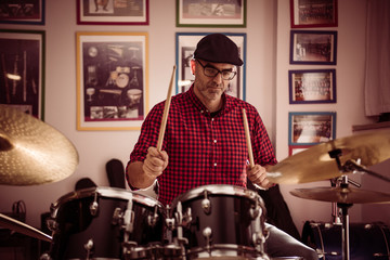 Fototapeta premium Caucasian young man with glasses and beret wearing a red and black plaid shirt, playing the drums with a soft side light