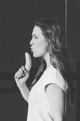 Fototapeta premium Black and white shot of Attractive young woman holding banana like gun on grey wall