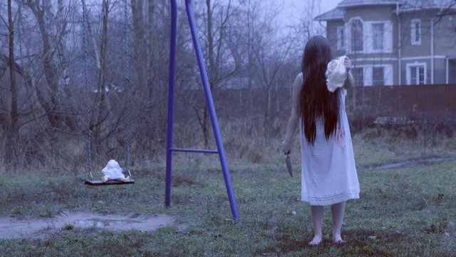 girl in a white dress with black long hair examines severed head of doll standing near swing.