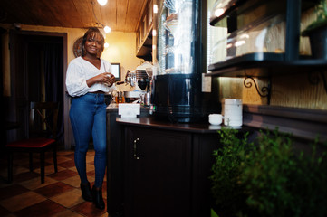 Stylish african american women in white blouse and blue jeans posed at cafe with caramel latte.