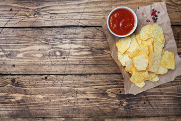 Spiced potato chips on paper and a plate of ketchup sauce on a wooden table. copy space.