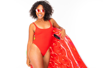 Pretty african female stands in swimsuit with rubber beach strawberry mattress and smiles isolated...