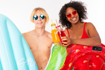 Pretty african female and caucasian blonde man stands in swimsuit with rubber beach mattresses, drinks juice and smiles isolated on white background