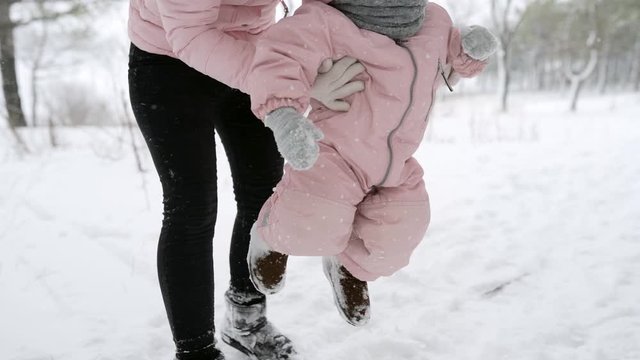Mother raises baby from the snow. Child in warm jumpsuit learns to walk. Girl walking doing first steps in winter forest under snowfall. Woman with daughter on snowy day outdoors. Warm pink romper.