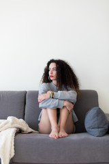 Obraz premium Pensive casual young woman sitting on a sofa at home and looking to the side.