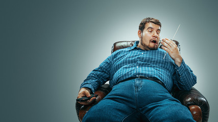 Obraz premium Portrait of fat caucasian man wearing jeanse and whirt sitting in a brown armchair isolated on gradient grey background. Emotional watching TV and changing channels, laughting. Overweight, carefree.