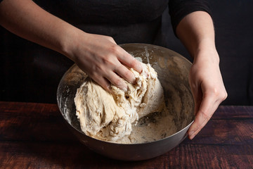 A cook in black kneads the dough in a metal bowl for cooking bread in the kitchen.