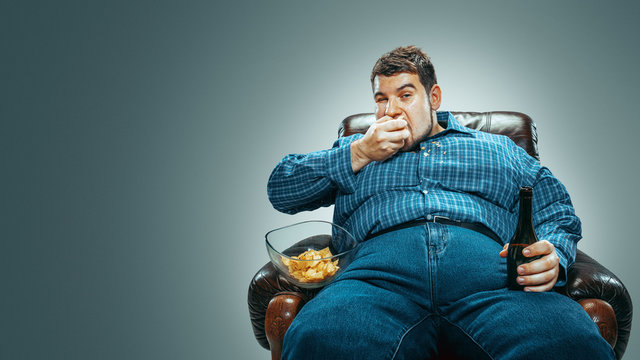 Portrait of fat caucasian man wearing jeanse and whirt sitting in a brown armchair isolated on gradient grey background. Watching TV drinks beer, eats chips and changing channels. Overweight, carefree