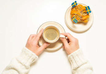 Swedish cinnamon bun (kanelbull) and coffee cup in hands on the white wooden table. Swedish flags. Coffee break (fika) concept. Top view.