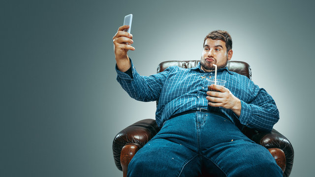Portrait of fat caucasian man wearing jeanse and whirt sitting in a brown armchair on gradient grey background. Making selfie with the cola, laughting. Overweight, carefree. Concept of weight loss.