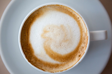 Close up white coffee cup with heart shape latte art foam on black wood table near window with light shade on tabletop at cafe.