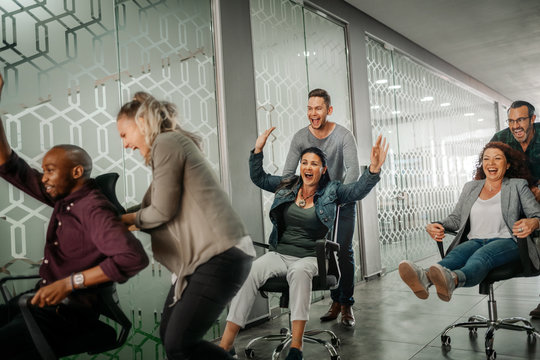 Cheerful business people in smart casual wear having fun while racing on office chairs and smiling
